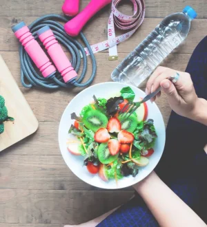 A person in workout clothes eating a healthy fruit and vegetable salad, surrounded by exercise equipment like a jump rope and dumbbells, illustrating the benefits of exercise and healthy eating.