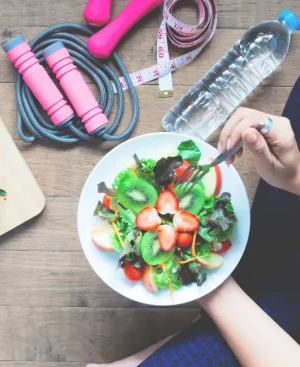 A person in workout clothes eating a healthy fruit and vegetable salad, surrounded by exercise equipment like a jump rope and dumbbells, illustrating the benefits of exercise and healthy eating.