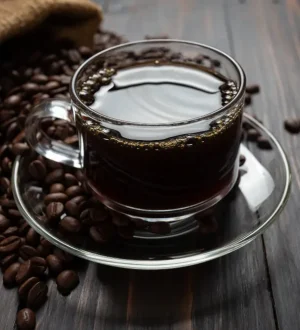 A clear glass cup of black coffee surrounded by beans on a wooden table, perfect to indulge your coffee cravings.
