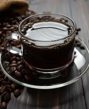 A clear glass cup of black coffee surrounded by beans on a wooden table, perfect to indulge your coffee cravings.