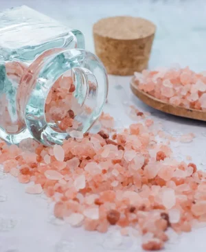Coarse Himalayan pink salt crystals spilling from a glass jar onto a table, representing sodium intake and blood pressure risks.