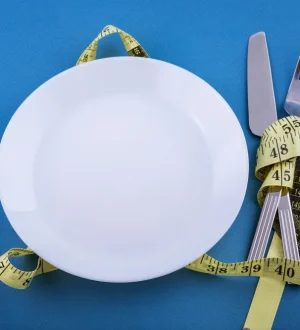 An empty white dinner plate and cutlery wrapped in a yellow measuring tape, representing the metabolic risks when you lose weight by starving yourself.