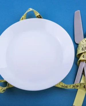 An empty white dinner plate and cutlery wrapped in a yellow measuring tape, representing the metabolic risks when you lose weight by starving yourself.