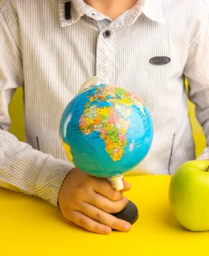 A person holding a small globe in one hand and a fresh green apple in the other over a bright yellow surface, symbolizing global health goals Managing Health Goals Across Time Zones.