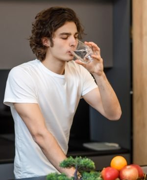A young man with curly hair drinking a glass of water in a modern kitchen to unlock weight loss.