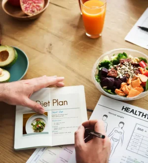 Top-down view of a doctor or nutritionist designing a personalized clinical diet plan on a wooden desk alongside a fresh vegetable salad bowl, sliced avocados, nuts, and a BMI weight chart.