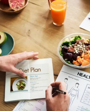 Top-down view of a doctor or nutritionist designing a personalized clinical diet plan on a wooden desk alongside a fresh vegetable salad bowl, sliced avocados, nuts, and a BMI weight chart.