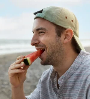 A smiling young man wearing a backward cap, eating a fresh slice of watermelon on a sunny beach, demonstrating healthy summer hydration and craving management.