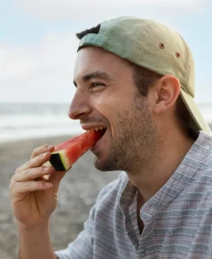 A smiling young man wearing a backward cap, eating a fresh slice of watermelon on a sunny beach, demonstrating healthy summer hydration and craving management.