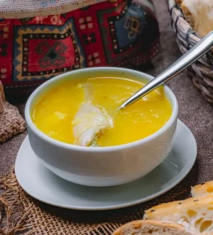A white bowl of golden chicken soup or shorba containing a chicken piece and a spoon, surrounded by traditional naans and bread baskets on a rustic brown fabric surface.