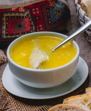 A white bowl of golden chicken soup or shorba containing a chicken piece and a spoon, surrounded by traditional naans and bread baskets on a rustic brown fabric surface.