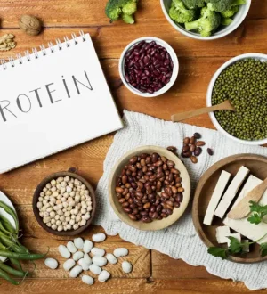 A flat-lay photograph of various vegan sources of protein, including kidney beans, lentils, chickpeas, tofu, nuts, and green beans, arranged on a wooden table around a notepad with the word 'PROTEIN'.