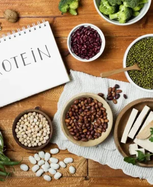 A flat-lay photograph of various vegan sources of protein, including kidney beans, lentils, chickpeas, tofu, nuts, and green beans, arranged on a wooden table around a notepad with the word 'PROTEIN'.