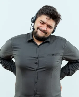 A man with a heavy build, wearing a grey shirt and a headset, grimaces in acute pain while holding his lower back with both hands.