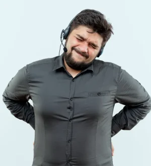 A man with a heavy build, wearing a grey shirt and a headset, grimaces in acute pain while holding his lower back with both hands.