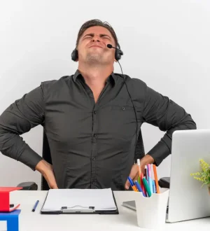 A male call center agent in Karachi, wearing a headset and wincing in pain while sitting at a desk, demonstrating the common suffering and need for non-surgical sciatica treatment.
