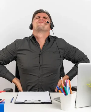 A male call center agent in Karachi, wearing a headset and wincing in pain while sitting at a desk, demonstrating the common suffering and need for non-surgical sciatica treatment.